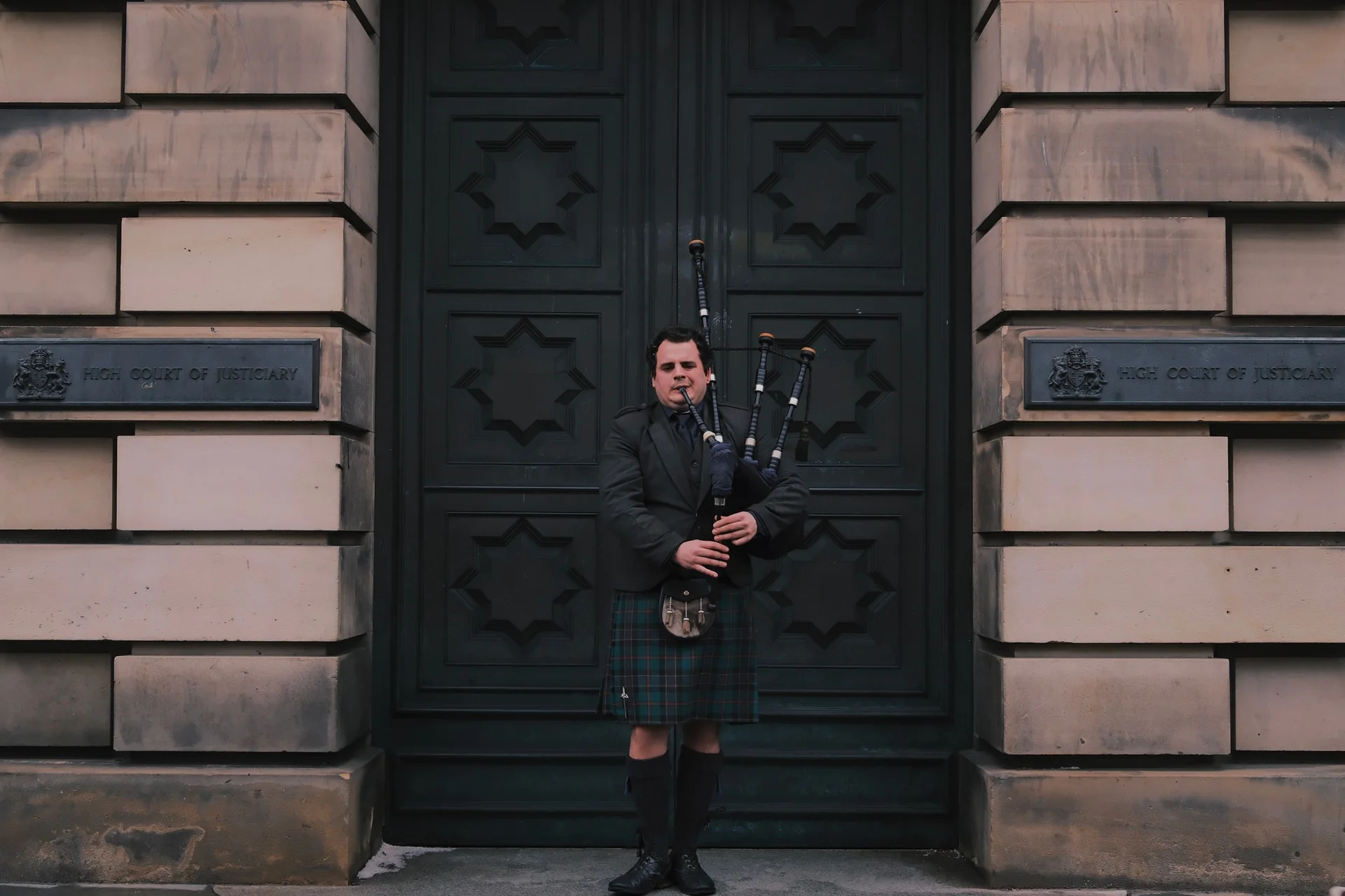 Dudák v plném skotském kroji hraje na dudy před honosným kamenným portálem v Edinburghu